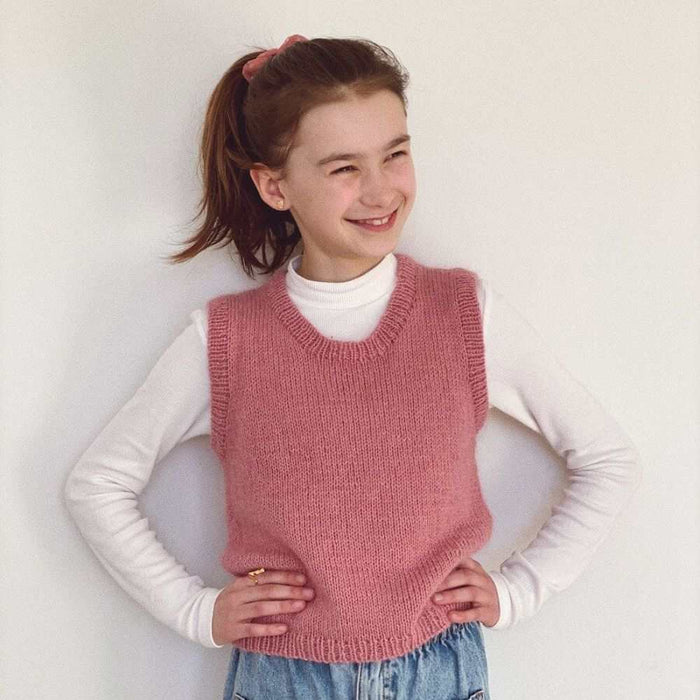 Child wearing pink Stockholm Slipover Junior vest by PetiteKnit, over a white long-sleeve shirt, smiling and posing with hands on hips.