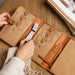 Leather circular needle knitting case in burned tan and gold, featuring multiple pockets, snap closures, and elastic band, ideal for organizing knitting accessories.