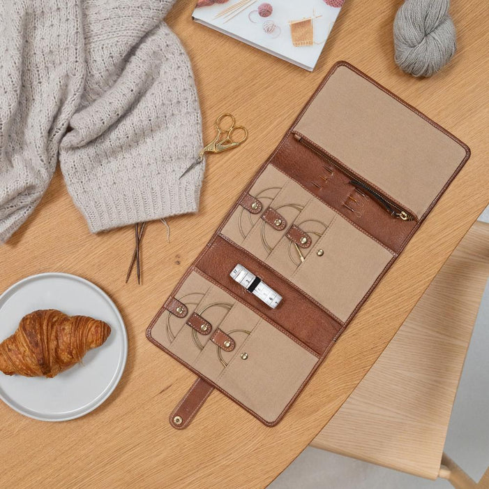 Knitting bag in walnut and gold leather with multiple compartments and snap closures, displayed on a wooden table.