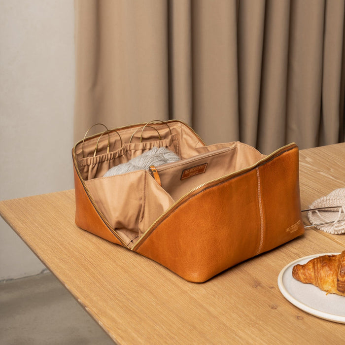 Knitting project bag in burned tan and gold leather with needle pockets on a wooden table.
