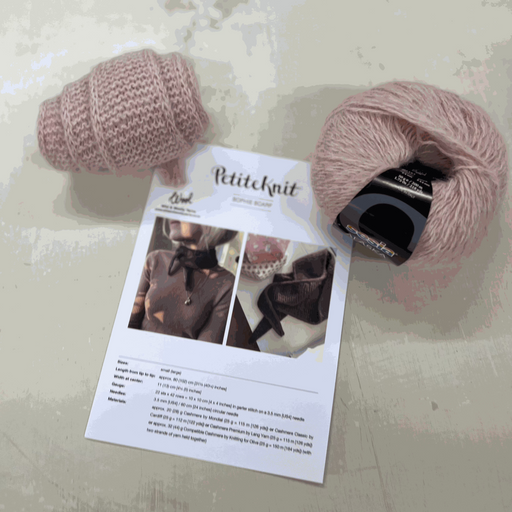 Sophie Scarf Knit Kit with pink yarn, pattern, and knitting supplies on a table.