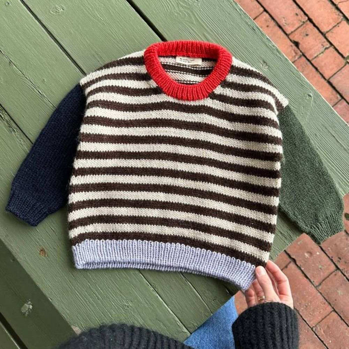 Holger Sweater Knitting Pattern by PetiteKnit displayed on a green wooden bench, showcasing striped body and contrasting colored sleeves and neck edge.