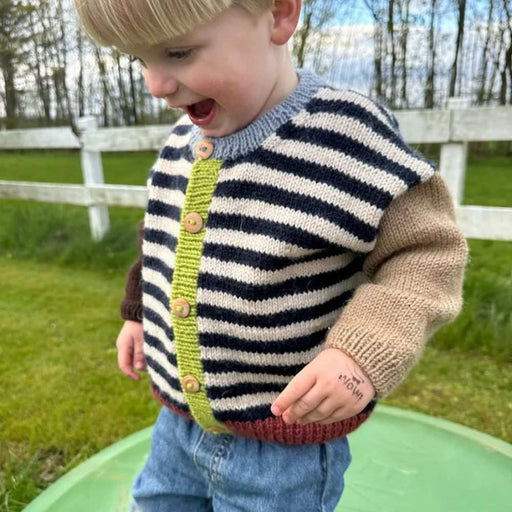 Child wearing handmade striped cardigan from PetiteKnit's Holger Cardigan knitting pattern, showcasing colorful design and button details outdoors.
