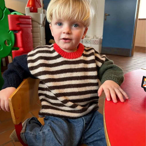 Child in a striped Holger Sweater, sitting on a chair, featuring different colored sleeves and a red neck edge, by PetiteKnit.