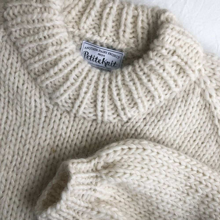 Holiday Sweater Knitting Pattern by PetiteKnit in cream wool with ribbed collar, showcasing texture and branding label.