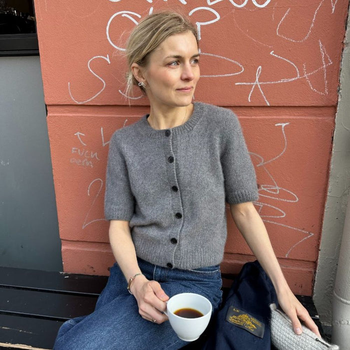 Woman wearing a gray short-sleeve Maggie Cardigan from PetiteKnit, sitting casually with a cup of coffee.