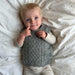 Baby wearing a gray cable-knit slipover, lying on a white blanket.