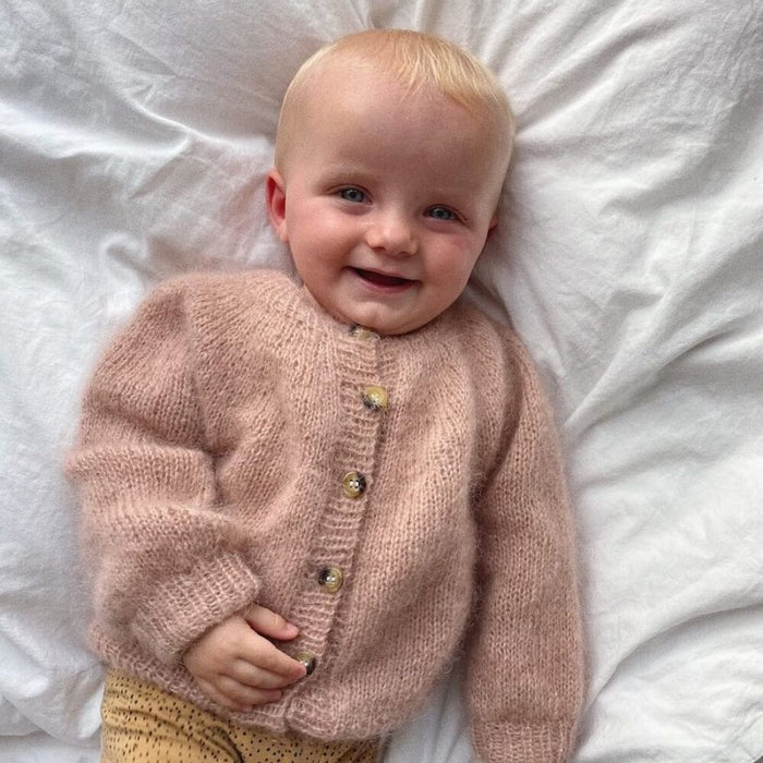 Baby wearing a hand-knitted mohair cardigan from PetiteKnit, showcasing the Novice Cardigan Mini design in soft pink with ribbed edges and buttons.
