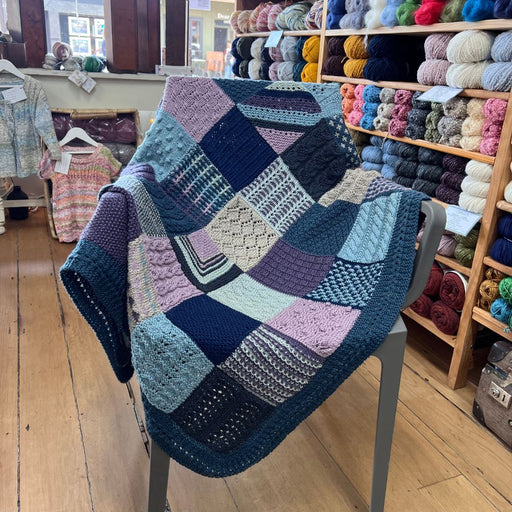 Sampler blanket knitting pattern on display, featuring 48 unique squares in various stitches and colors, completed with a knitted border.