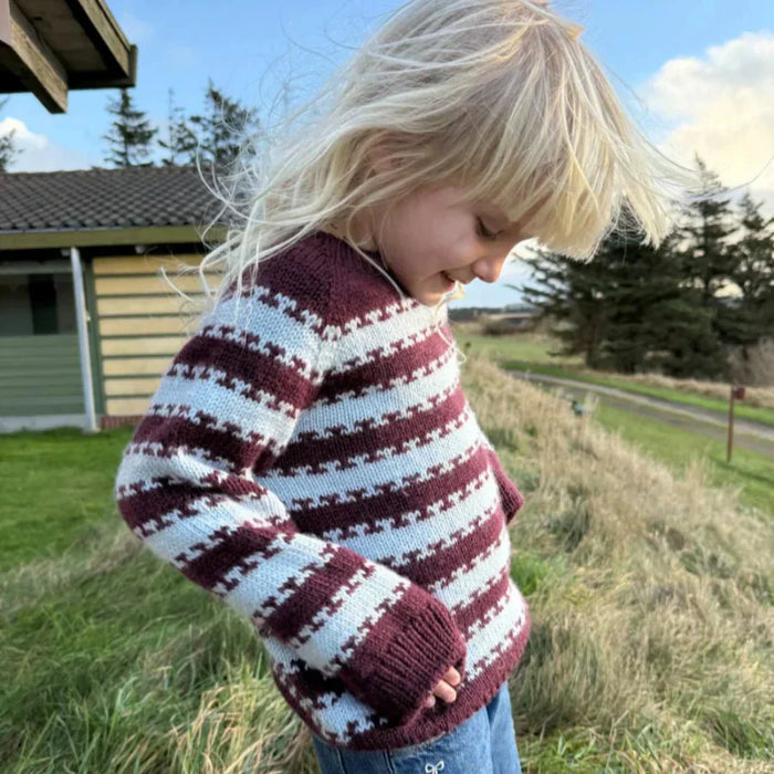 Child wearing a striped knit sweater outdoors, showcasing the "Key Sweater Junior" pattern by PetiteKnit.