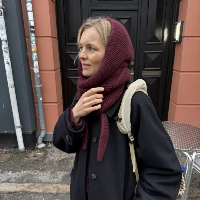 Cozy woman wearing maroon Sophie Hood scarf, showcasing elegant knitting pattern.
