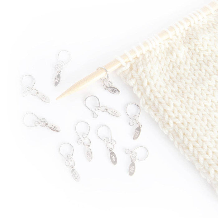 Silver cast on counting stitch markers numbered in increments of 50 with easy-to-secure melody clips, shown with a knitted white fabric and wooden knitting needle.