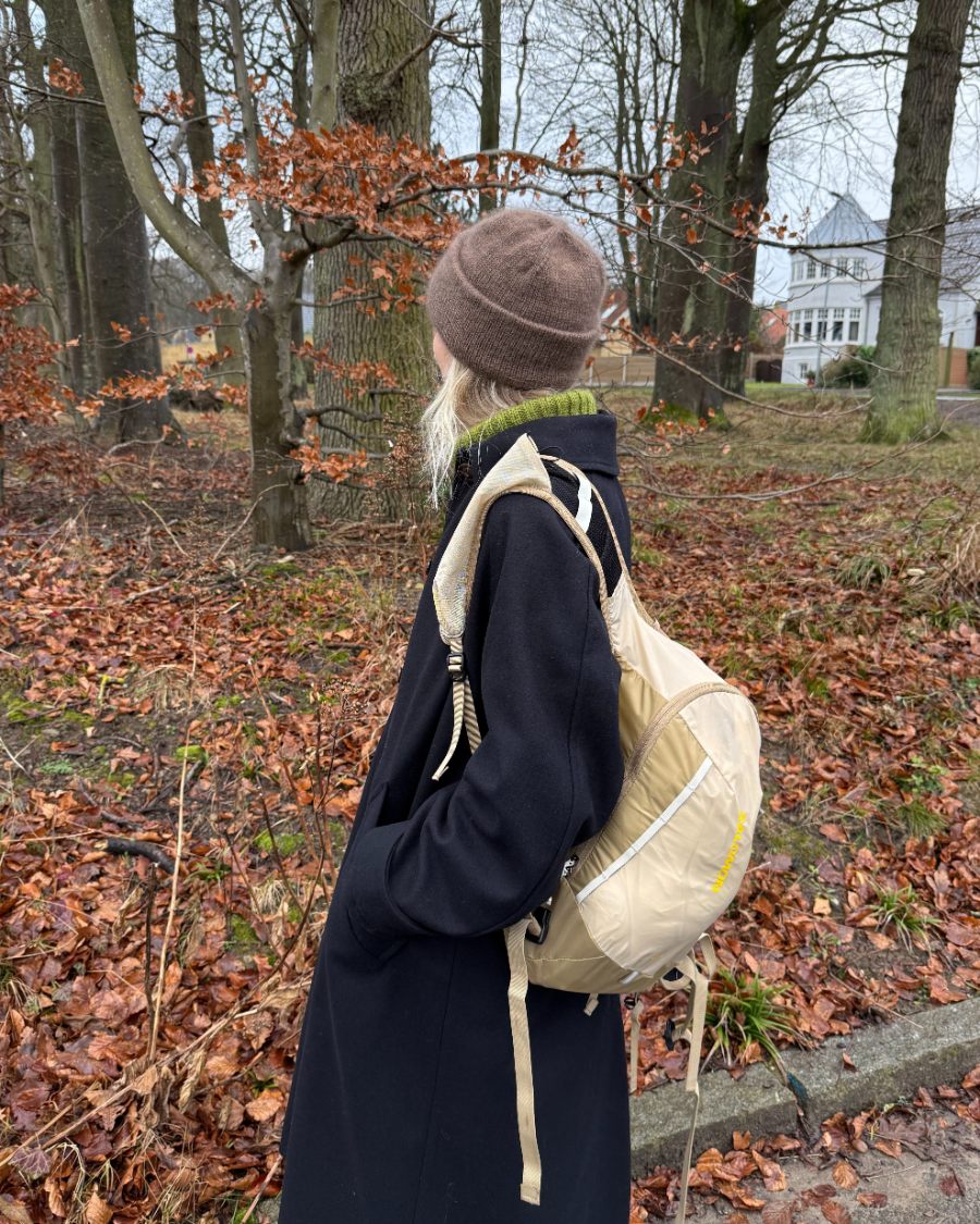 Oslo Hat by PetiteKnit: brown knitted hat worn with a dark coat in an autumn garden
