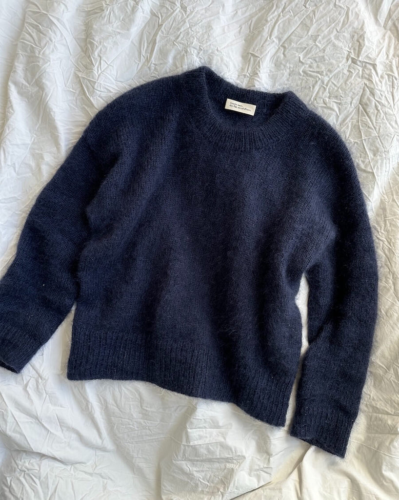 Navy blue Stockholm Sweater by PetiteKnit laid flat on white bedding, showing the round neckline, long sleeves and ribbed edges