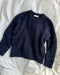 Navy blue Stockholm Sweater by PetiteKnit laid flat on white bedding, showing the round neckline, long sleeves and ribbed edges